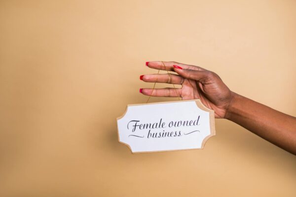 Close-up of a woman's hand holding a 'Female Owned Business' sign on a beige background.