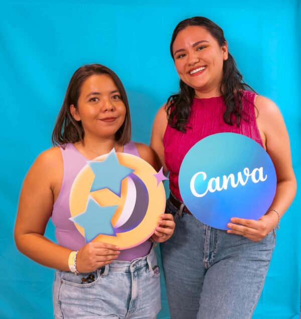 Two women smiling and holding a Canva sign against a blue background, promoting creativity and design.